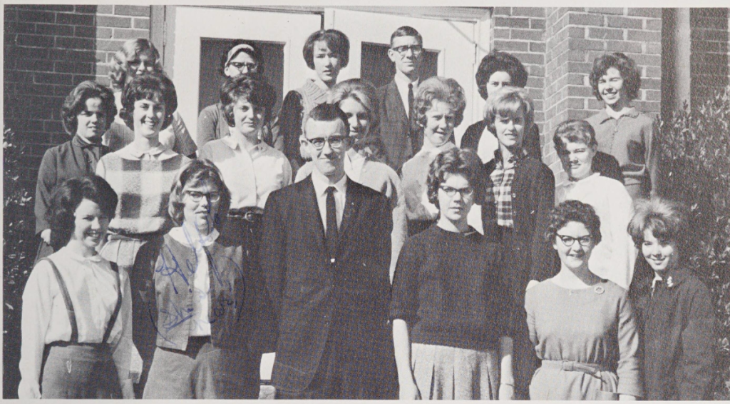 A smiling group of nineteen high school students stand in front of a brick building. These students are the members of the library club.
